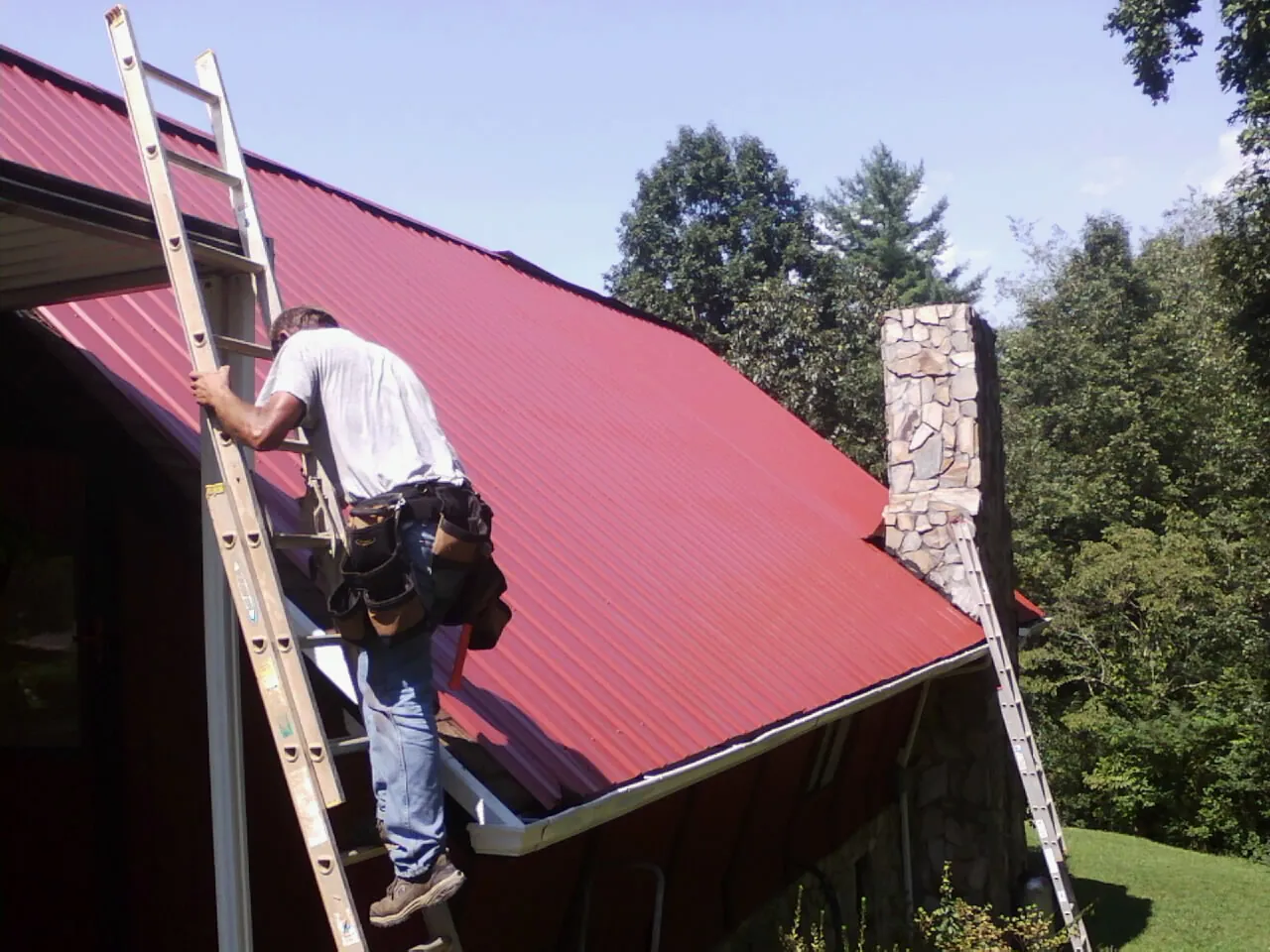 Roofer on a ladder beside a red metal roof and stone chimney — Soffit & Fascia Repair by Roofing Brothers in Ennice